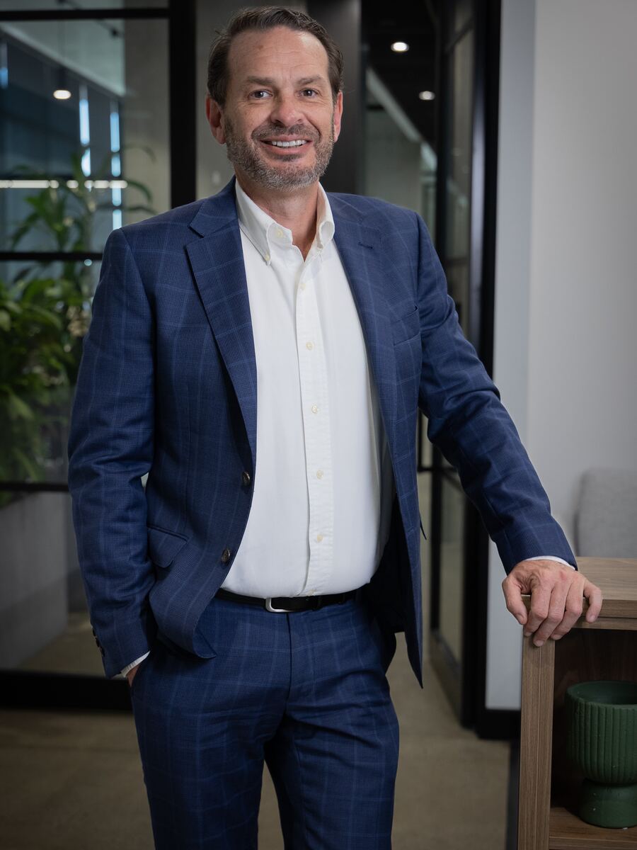 Cameron, Sparrow Advisory director, smiling in a navy checked suit and open-collar white shirt, standing in the Brisbane office
